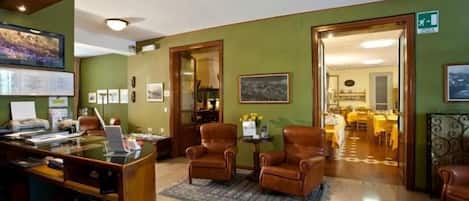 Un hall de hotel con mostrador de recepción, sillones de cuero, zona de comedor y una escalera que conduce a los pisos superiores.
