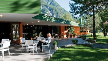 Un edificio con una zona salotto esterna coperta, una persona che legge un giornale e un paesaggio montano sullo sfondo.