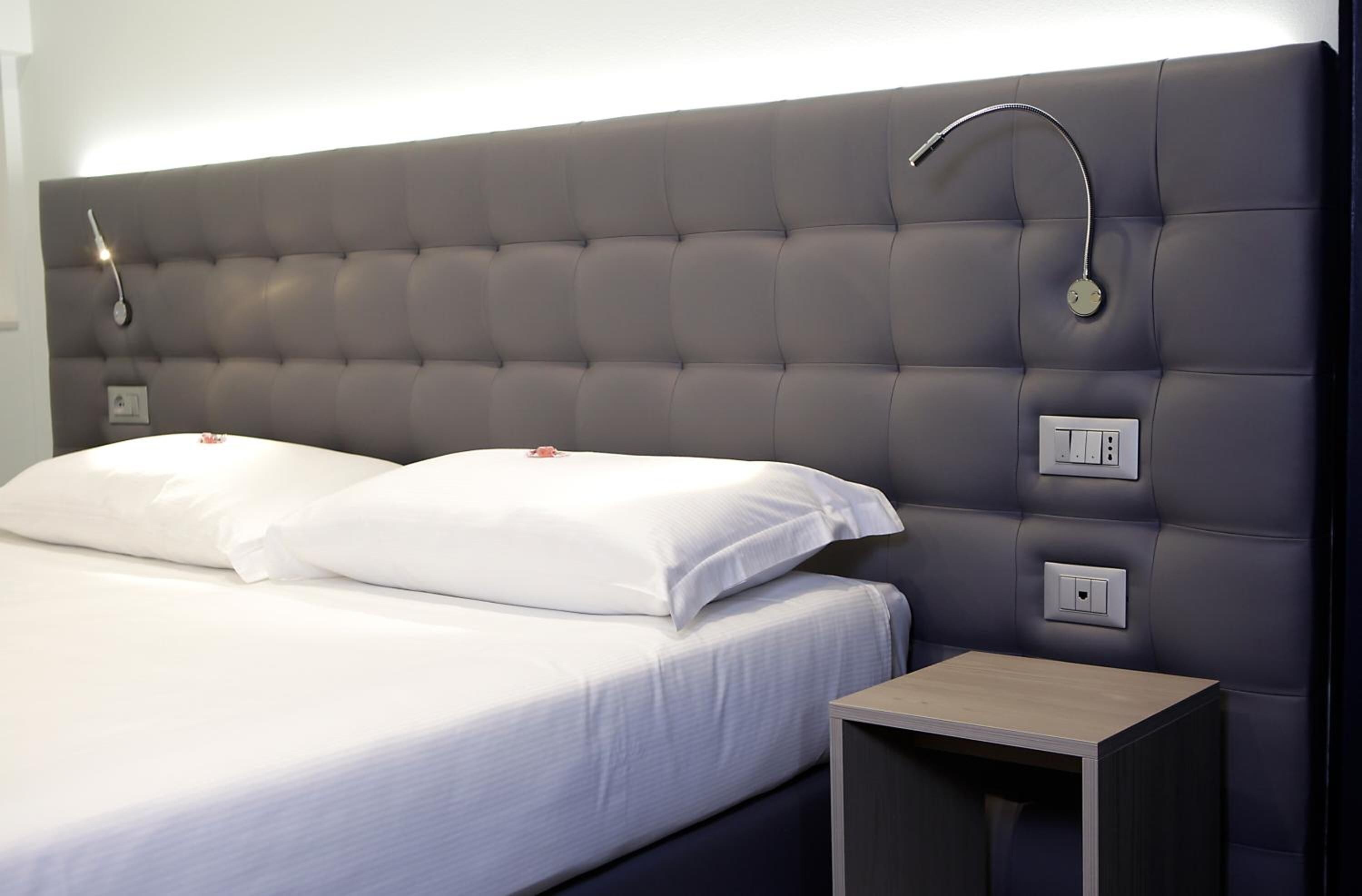 Un letto rifatto con lenzuola bianche e due cuscini, un comodino con una lampada e un punto luce a parete.