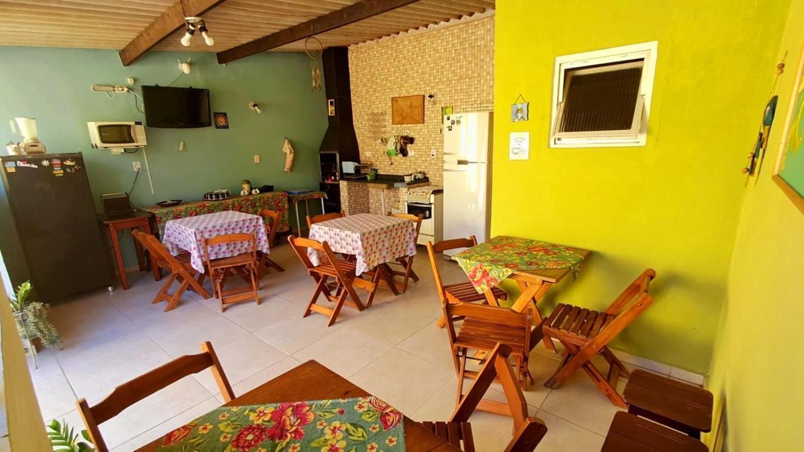 A dining area with wooden tables and chairs, a kitchen in the background, and a bright yellow wall.