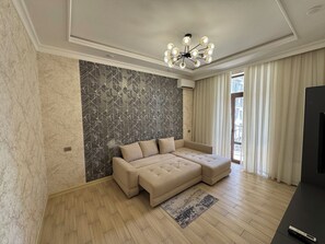 Un salon moderne avec un canapé d’angle, un papier peint à motifs, un lustre et une grande fenêtre avec des rideaux.