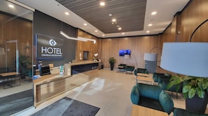 Zona de recepción de hotel con diseño moderno, que incluye un mostrador de recepción, área de descanso y una pantalla de televisión.