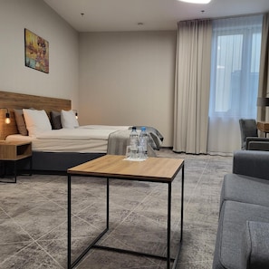 A hotel room with a bed, a small table with water bottles, a chair, and a painting on the wall.