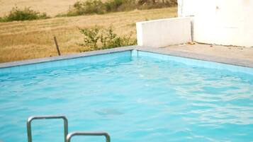 Una piscina con una escalera metálica, rodeada por un suelo de cubierta verde y una pared blanca.