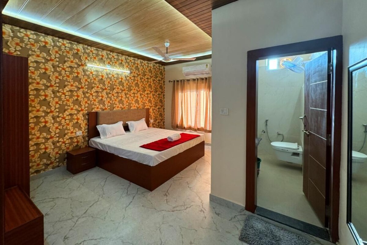Une chambre à coucher avec un lit, une tête de lit en bois, une table de chevet et un mur orné d’un motif floral. On peut apercevoir la salle de bain à travers une porte ouverte.