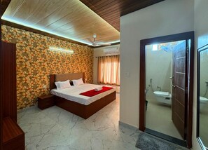 Une chambre à coucher avec un lit, une tête de lit en bois, une table de chevet et un mur orné d’un motif floral. On peut apercevoir la salle de bain à travers une porte ouverte.