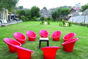 Un espace extérieur verdoyant, avec des chaises rouges disposées en demi-cercle, une petite table et, au fond, des tables vertes.