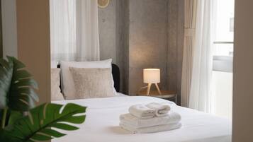 Un lit bien fait, avec du linge de lit blanc et une pile de serviettes pliées, une lampe de chevet et une plante au premier plan.