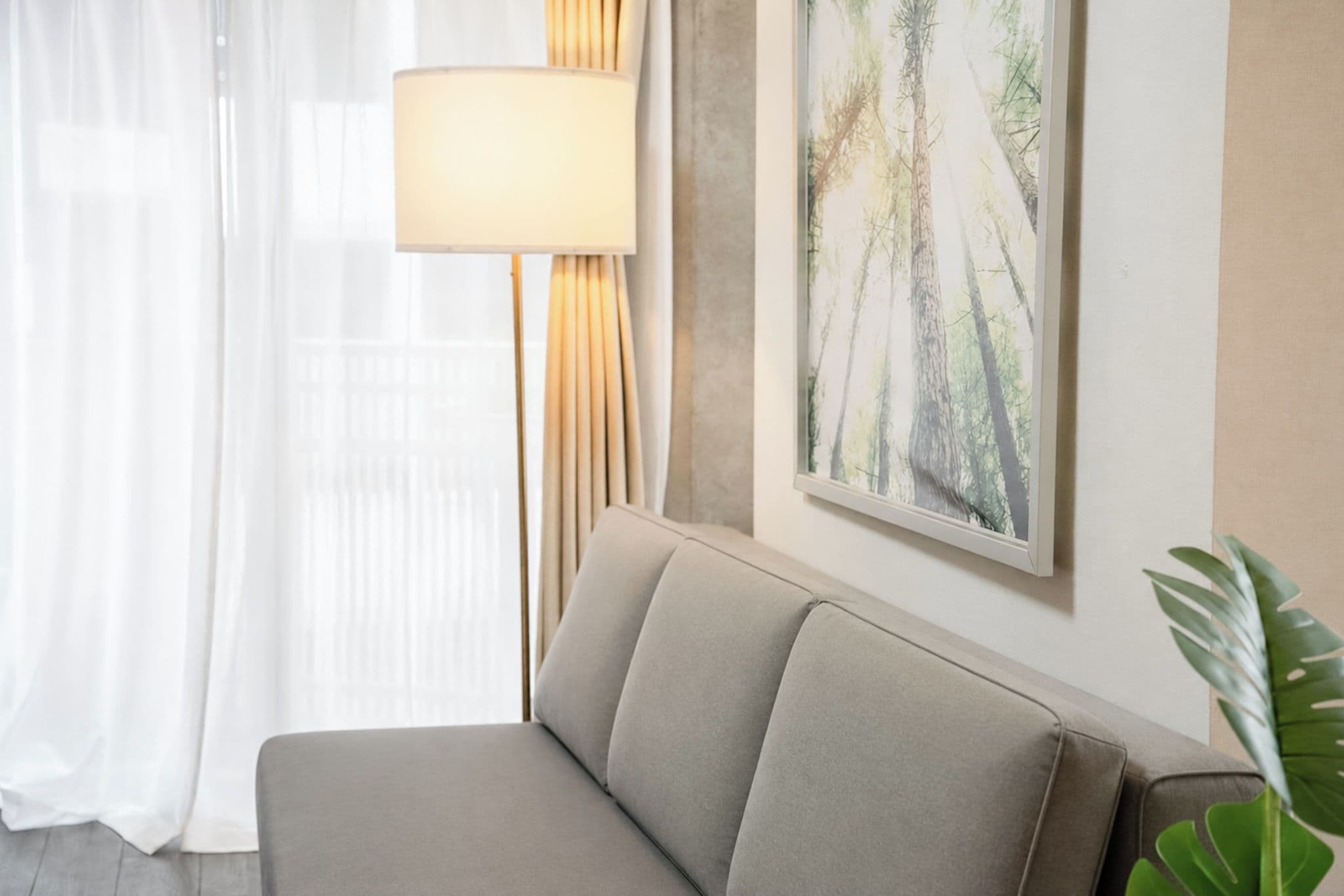 A neutral-colored sofa in a room with a large window, a floor lamp, and a framed nature picture on the wall.
