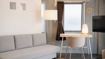 A modern living room with a grey sofa, a wooden desk with a chair, a television, and a window with curtains.