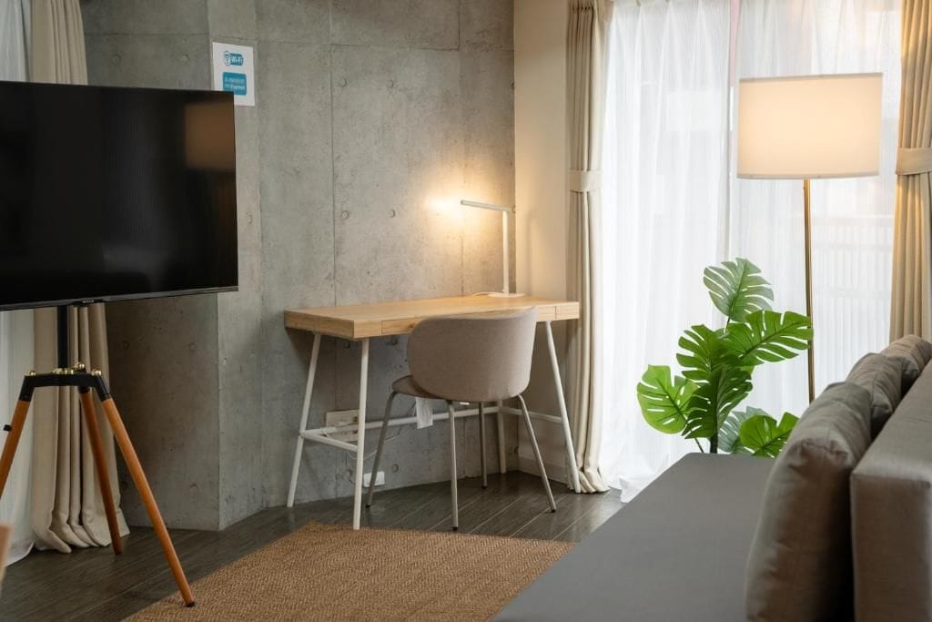 Une chambre moderne équipée d’un téléviseur à écran plat, d’un bureau avec une chaise, d’une lampe, d’un canapé et d’une grande plante.