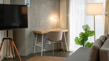 Une chambre moderne équipée d’un téléviseur à écran plat, d’un bureau avec une chaise, d’une lampe, d’un canapé et d’une grande plante.