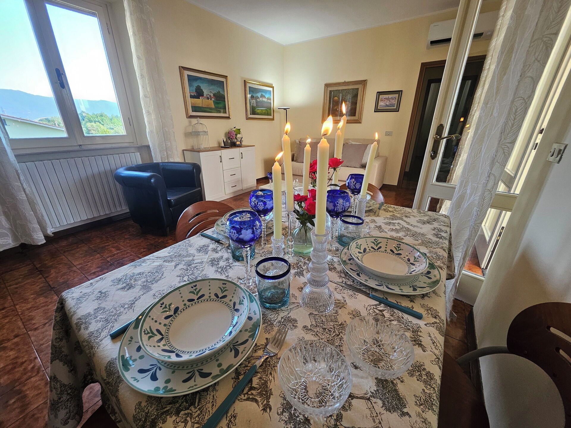 Uma sala de jantar com uma mesa posta para quatro pessoas, com toalhas de mesa estampadas, copos de vidro azuis e velas brancas.
