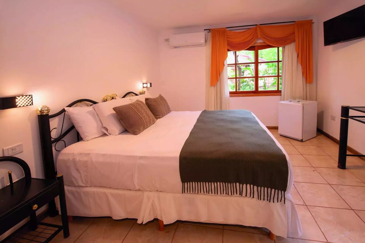 A bedroom with a bed, bedside table, lamp, window with curtains, and a TV mounted on the wall.