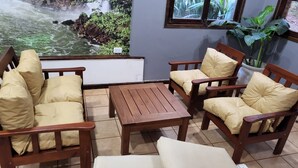 Un'area salotto con mobili in legno e cuscini, pavimento piastrellato e vista su una cascata attraverso una finestra.