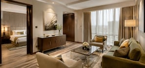 Quarto de hotel com cama, área de estar com sofá e poltronas, mesa de centro, armário e uma janela grande com cortinas.