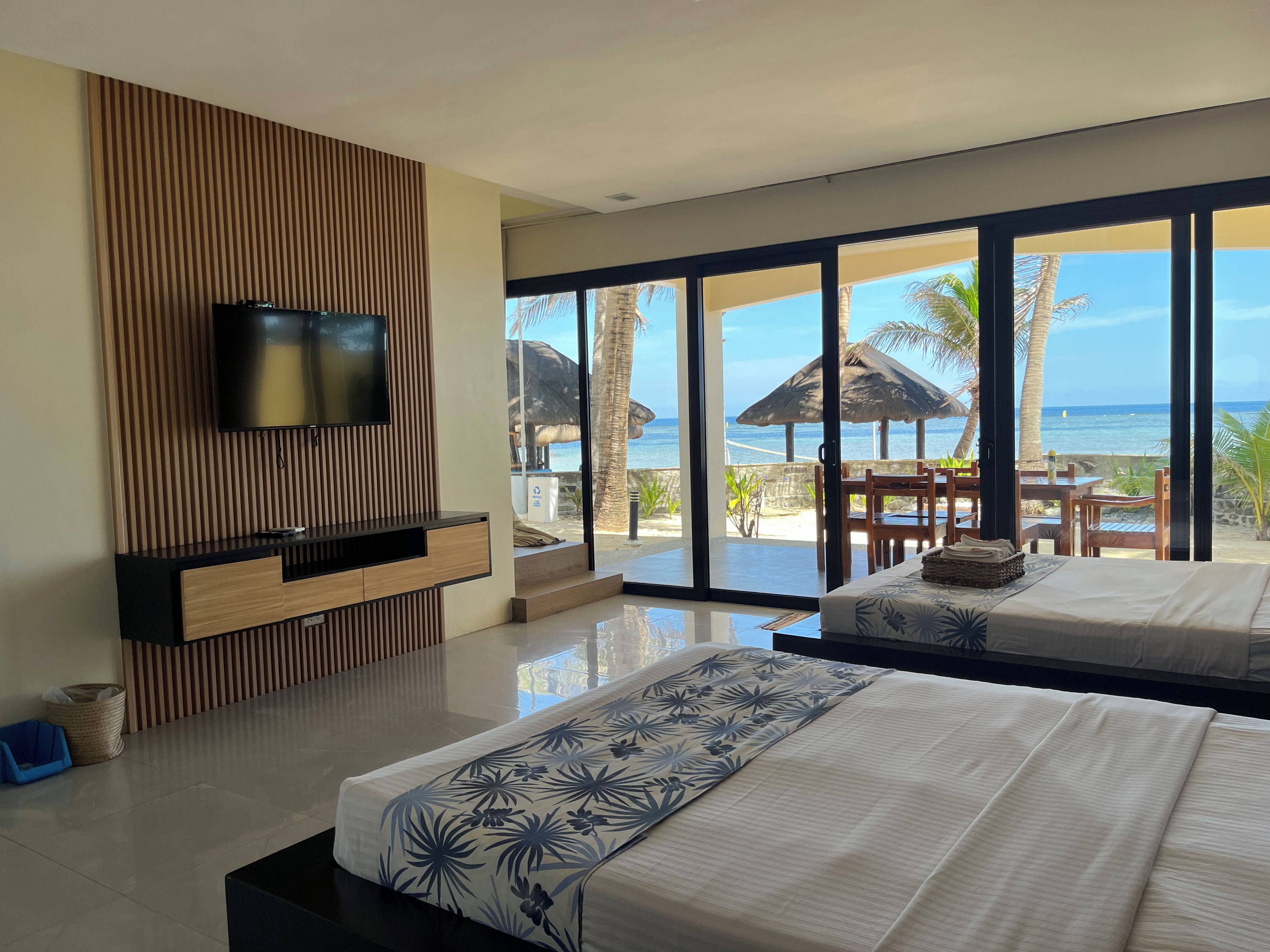 Habitación de hotel moderna con una cama grande, un televisor montado en un panel de madera y vista a la playa a través de puertas de vidrio amplias.