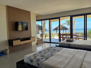 A modern hotel room with a large bed, a TV mounted on a wooden panel, and a view of the beach through large glass doors.
