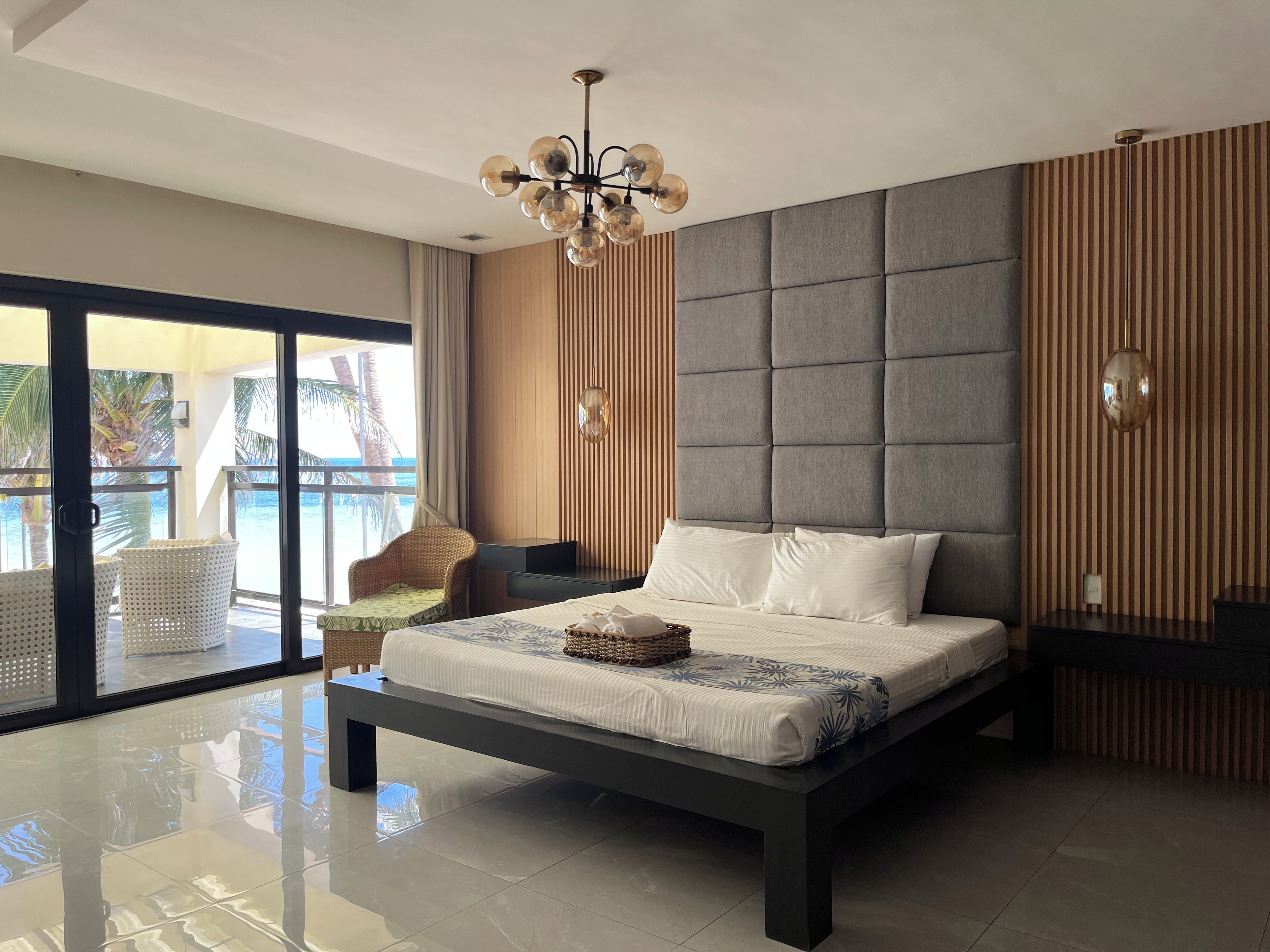 A modern bedroom with a large bed, a chandelier, a view of the ocean, and a balcony with wicker chairs.