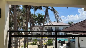 Vista desde un balcón con piscina, palmeras y un edificio blanco con balcones.