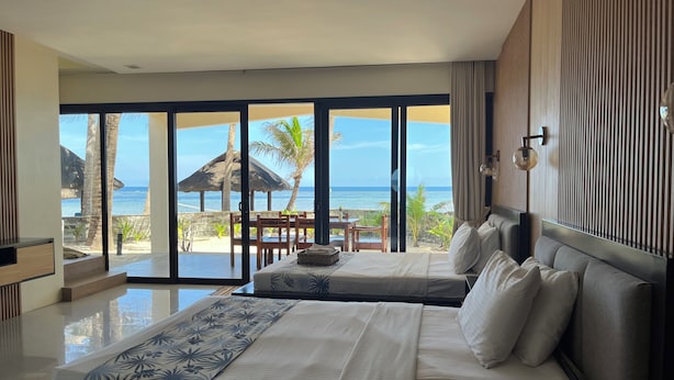 Habitación de hotel con una cama grande, vista a la playa y zona de comedor con mesa y sillas.