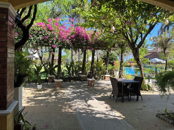 An outdoor patio area with a table and chairs, surrounded by potted plants and trees with pink flowers.