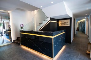 Una zona de recepción con mostrador de mármol, barandillas de vidrio, una escalera y una pared con el nombre 'IL BELVEDERE'.