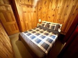 Quarto com uma cama, paredes de madeira, uma porta e uma mesa de cabeceira com abajur.