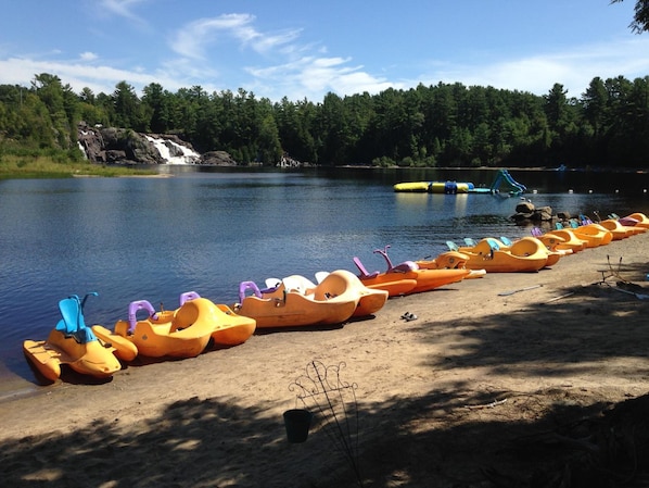 Una hilera de botes amarillos se alinean en una orilla arenosa junto a un lago tranquilo, con un área boscosa y una cascada al fondo.