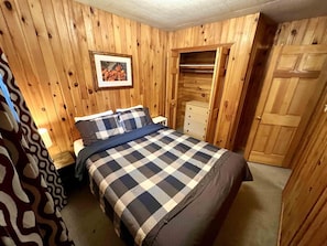 Un dormitorio con un revestimiento de madera, una cama con colcha a cuadros, un aparador y un armario.