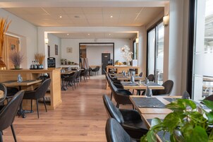 Restaurante com mesas e cadeiras de madeira, uma passagem central e vista para a rua através de grandes janelas.