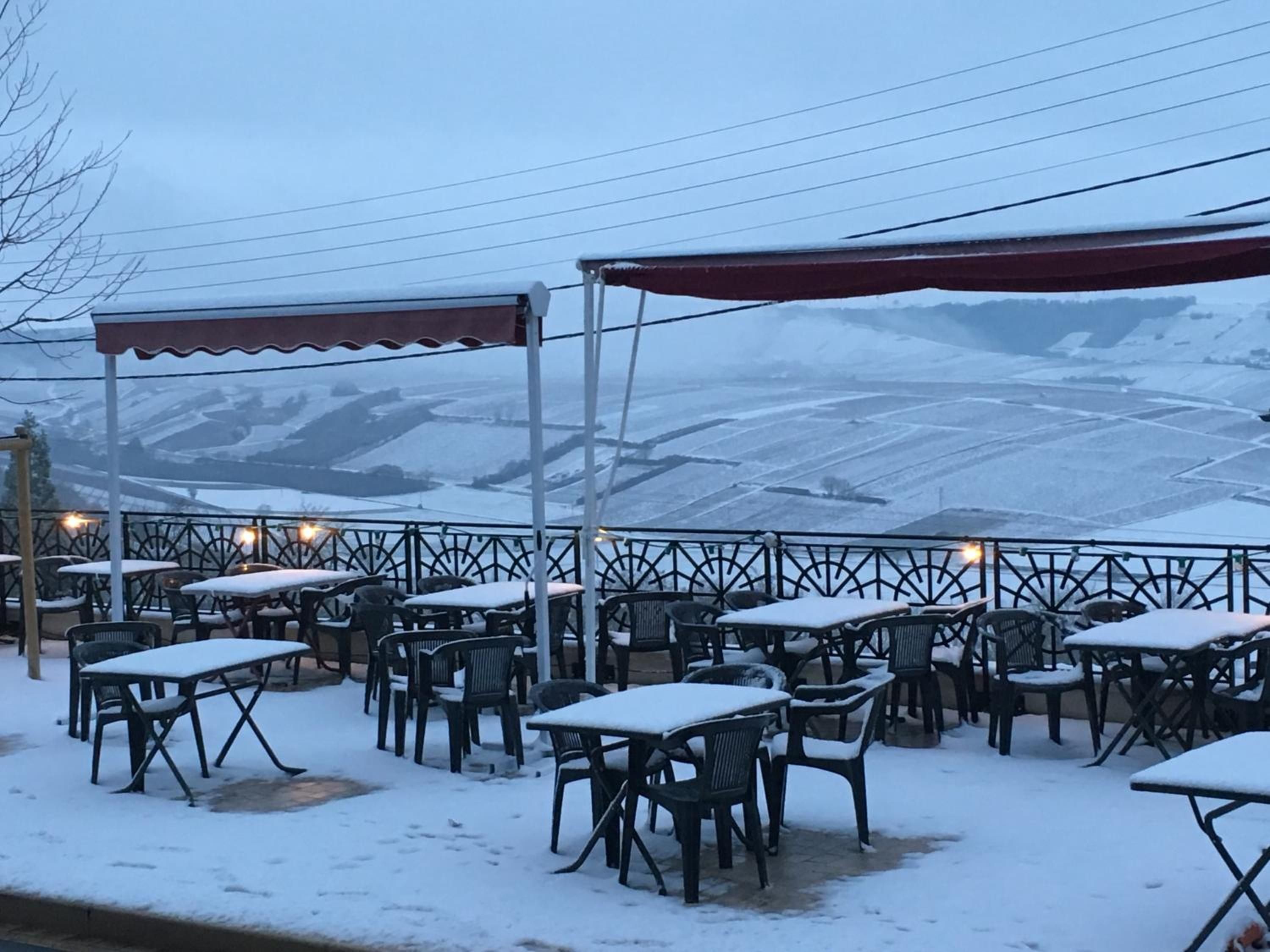 Ein Sitzbereich im Freien mit Tischen und Stühlen, die von Schnee bedeckt sind, einer roten Markise und Blick auf verschneite Felder.
