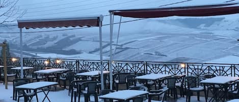 An outdoor seating area with tables and chairs covered in snow, a red awning, and a view of snow-covered fields.
