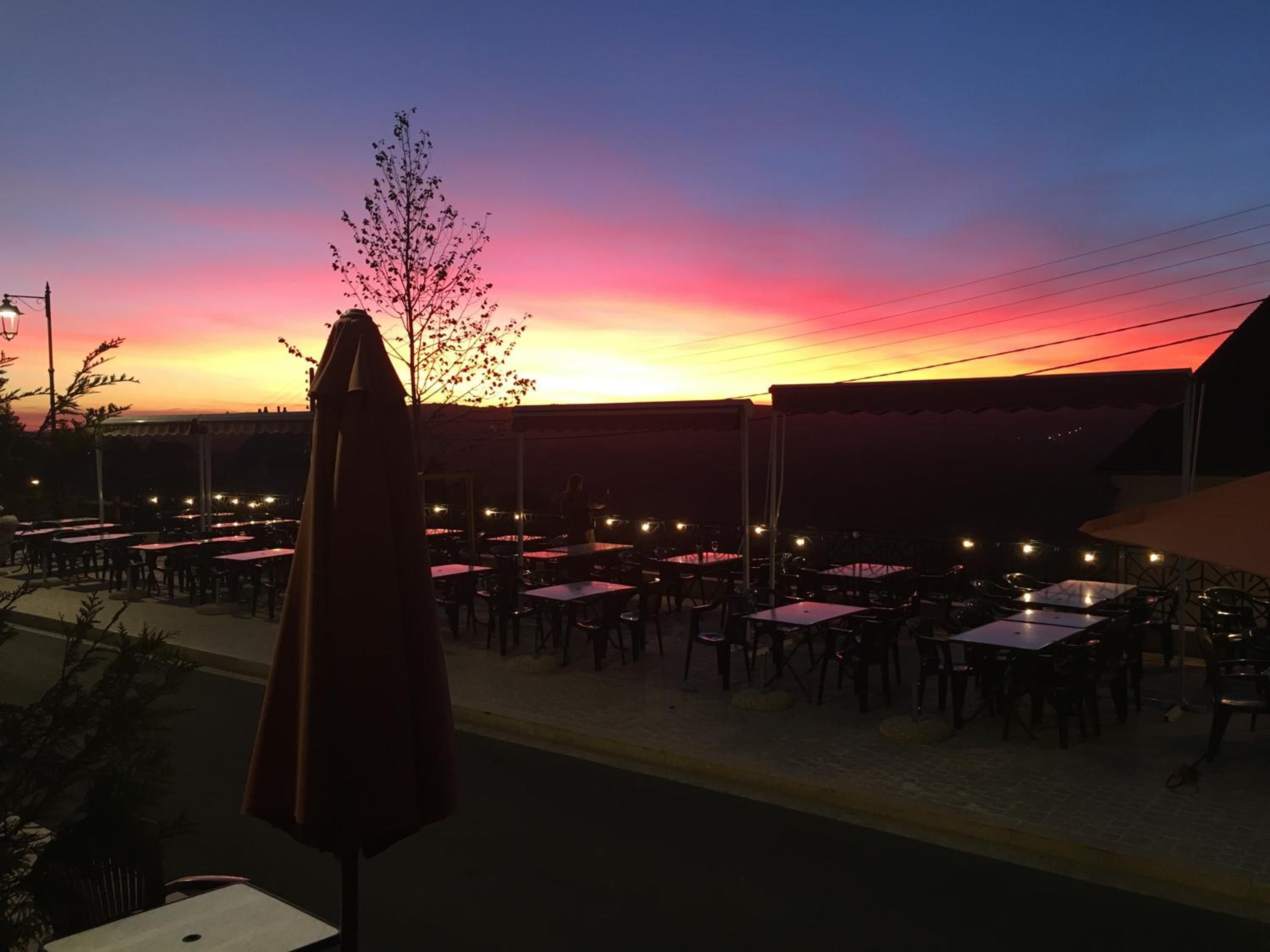 Een terras met tafels en stoelen, een boom en een zonsondergang op de achtergrond.