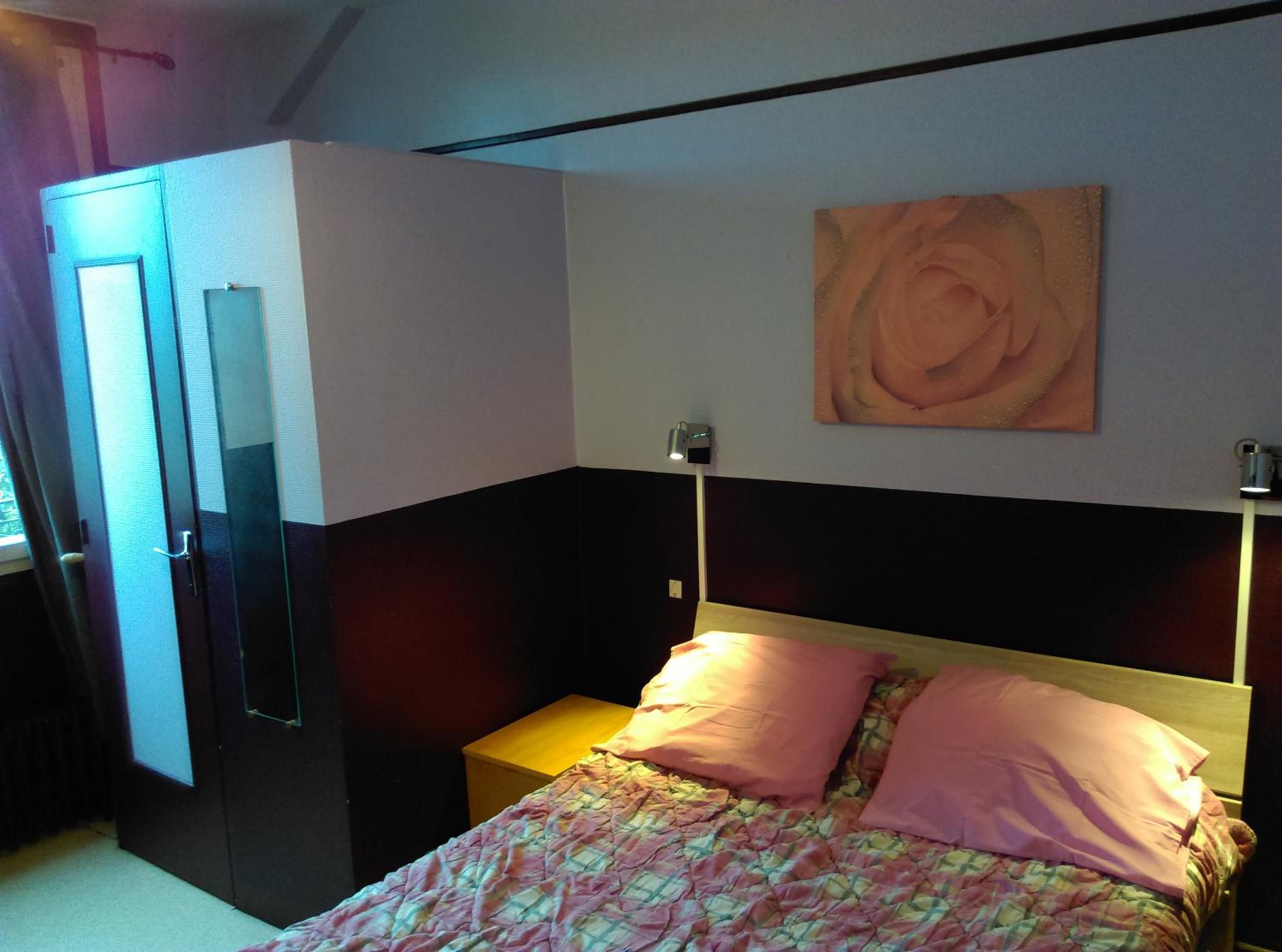 A bedroom with a bed, a nightstand, a wall-mounted lamp, and a door.