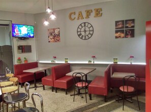 Interior de una cafetería con mesas y sillas, y bancos rojos, un reloj y un televisor en la pared.