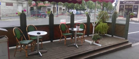 An outdoor seating area with wicker chairs and tables, a covered awning, and a view of the street with parked cars and buildings.