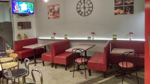 Interior de café com assentos em vermelho, mesas e cadeiras, um relógio e uma televisão na parede.