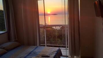 Quarto com cama, cortinas e vista do pôr do sol sobre a água.