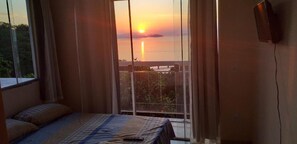Quarto com cama, cortinas e vista do pôr do sol sobre a água.