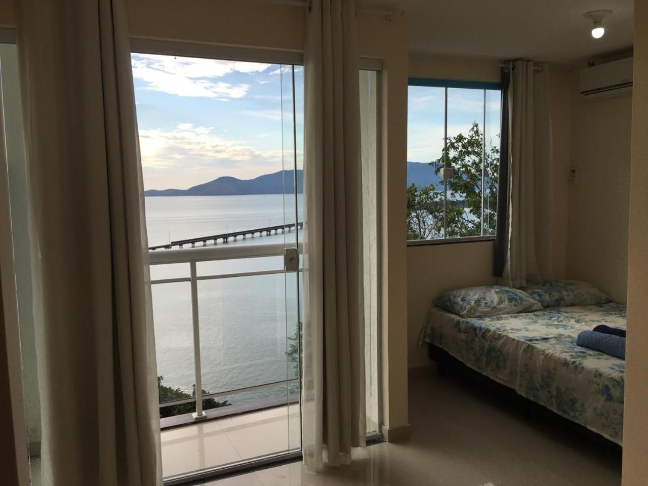 Quarto com cama, janela com vista para um corpo d'água e montanhas, e varanda com guarda-corpo.