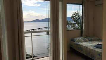 Quarto com cama, janela com vista para um corpo d'água e montanhas, e varanda com guarda-corpo.