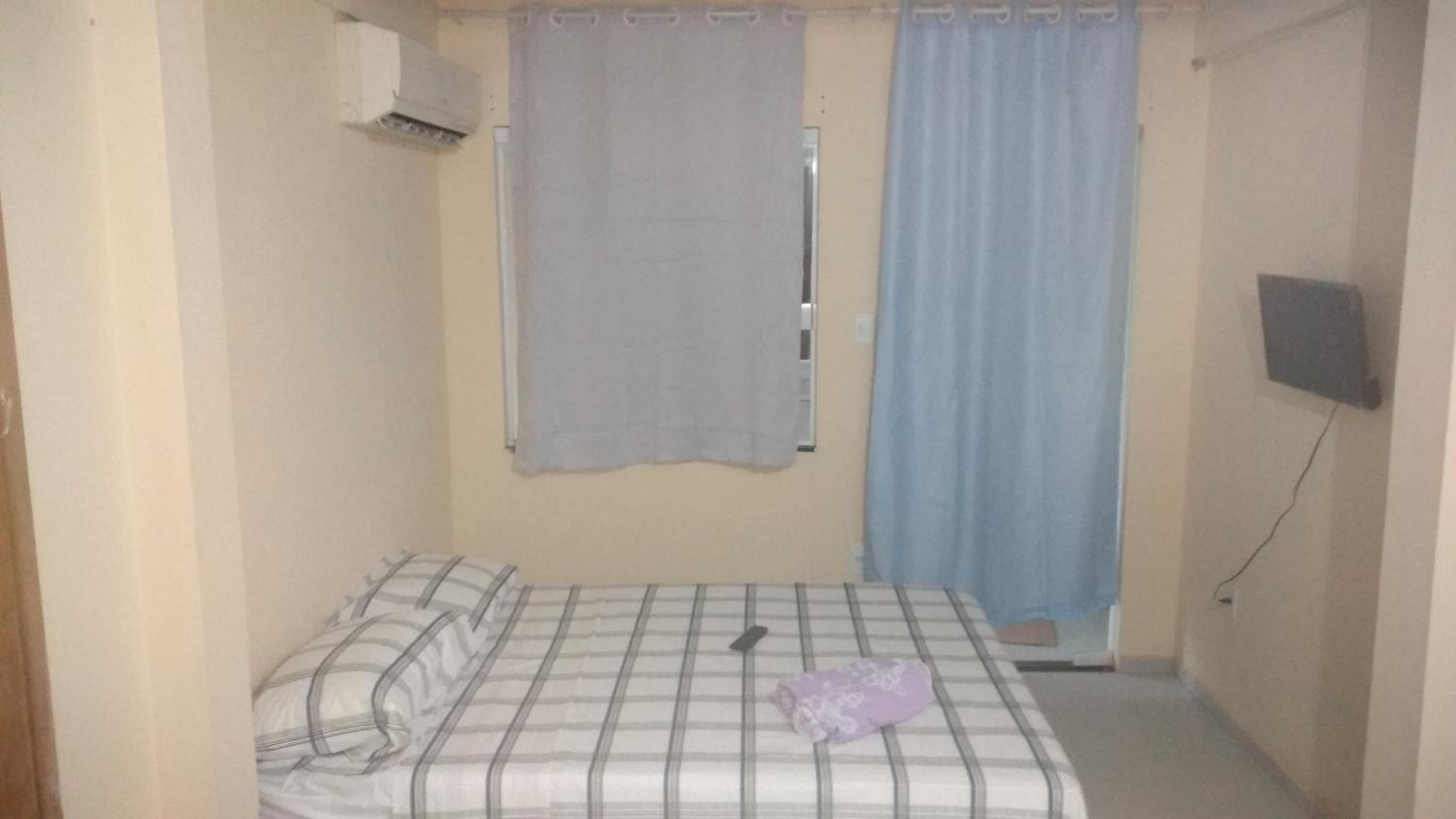 A small, single-bed room with a checkered bedspread, a pillow, a blanket, and a wall-mounted TV.