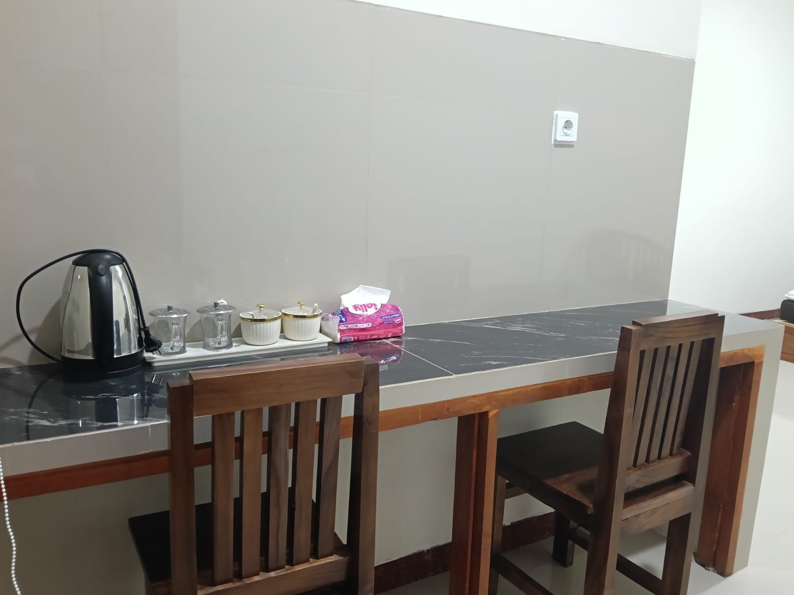 Un coin repas avec une table carrelée en noir et blanc, deux chaises en bois, une bouilloire, des récipients en verre et une boîte à mouchoirs rose.