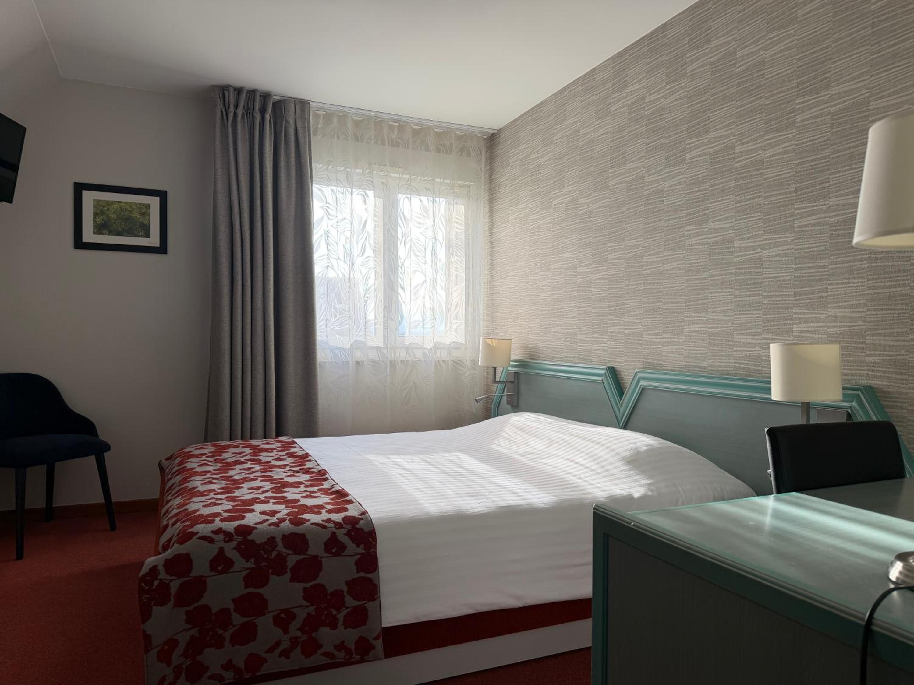 Hotellihuone, jossa on sänky, työpöytä, tuoli, ikkuna verhoilla ja kuvioitu seinä.