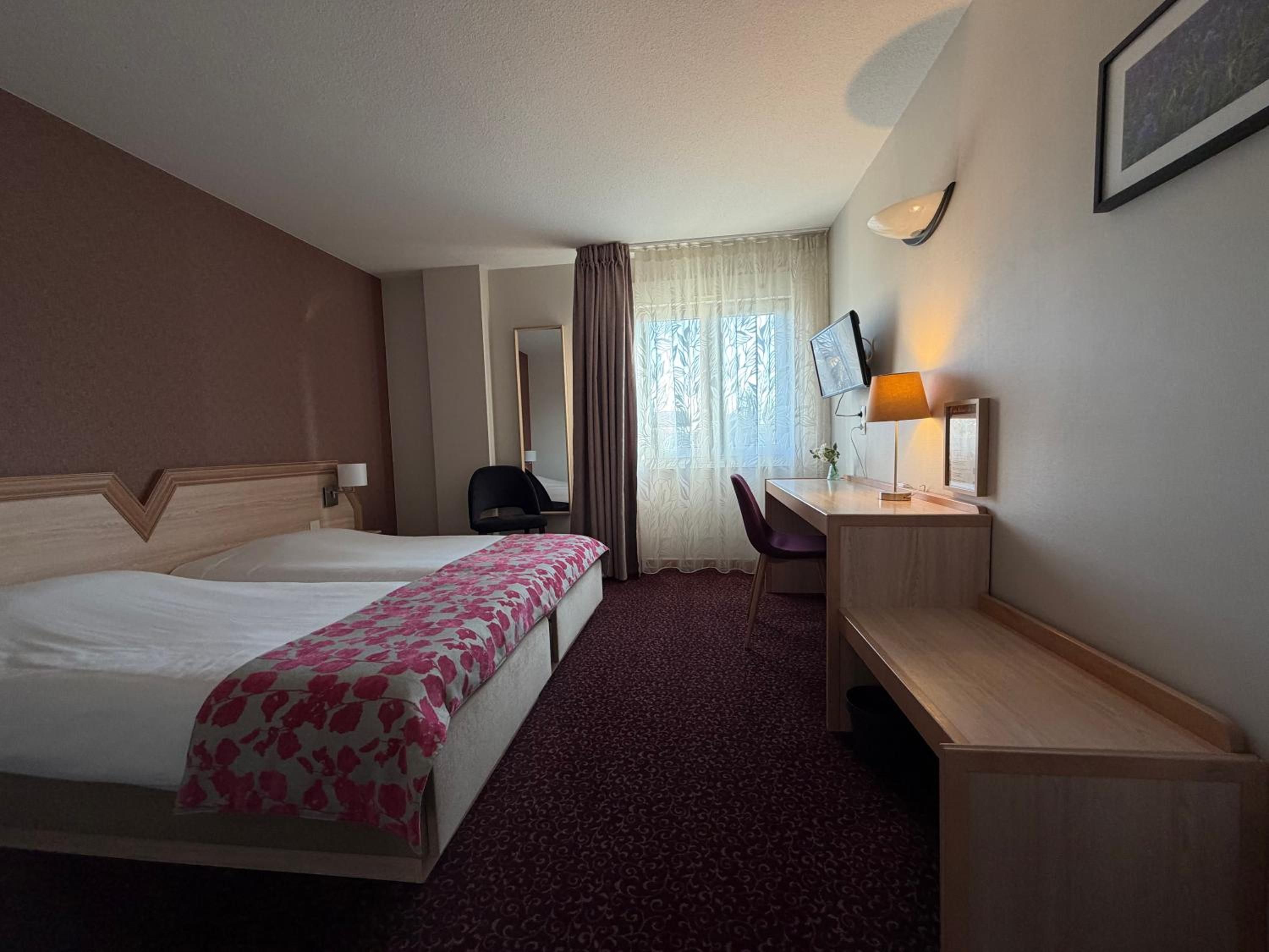 Une chambre d’hôtel avec deux lits, un bureau, une chaise, une télévision et une fenêtre avec des rideaux.