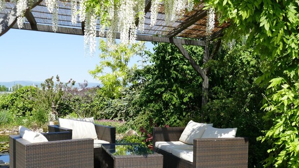 An outdoor seating area with wicker furniture and a glass table, surrounded by lush greenery and a pergola with hanging flowers. - Au Relais De L'ill (Riom)
