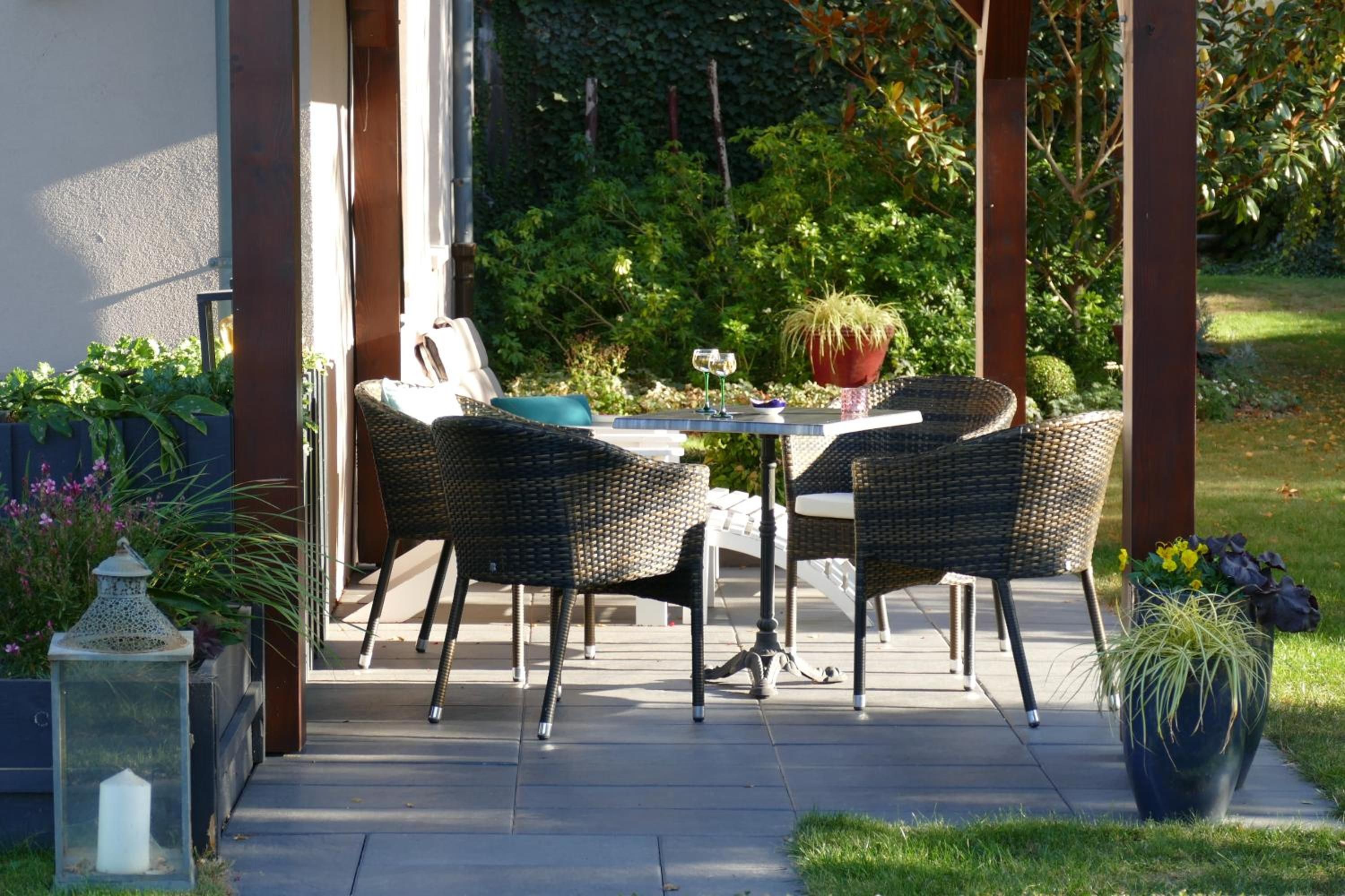 An outdoor patio with a table and two wicker chairs, surrounded by greenery and a white fence.
