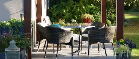 An outdoor patio with a table and two wicker chairs, surrounded by greenery and a white fence.