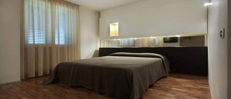 A hotel room with a bed, curtains, a flat-screen TV, and a wooden floor.
